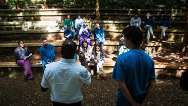 A group of people sit on shaded outdoor seating listening to two speakers