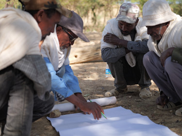 A group of four people crouches over a sheet of paper on the ground and one of them writes on the paper