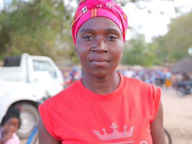 A woman in a pink headscarf looks at the camera with a tree and street behind her