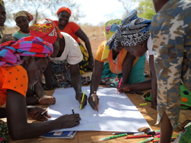 A group of people sit on the floor and write on a big sheet of paper