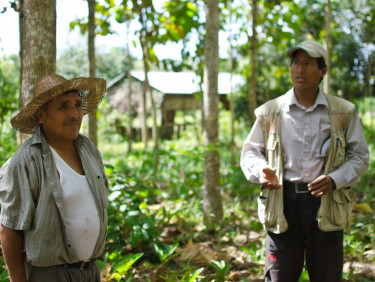 Two men in hats stand talking in a wood with a house in the background