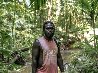 A man in a pink sleeveless t-shirt stands on a path in a forest