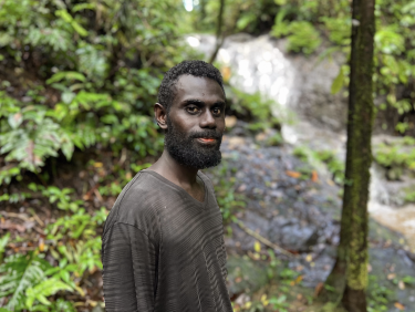 A man in a grey shirt stands in a forest in front of a small waterfall