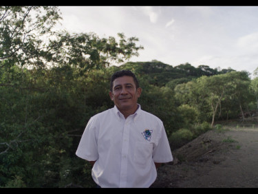 A man in a white shirt stands on a path with trees on one side