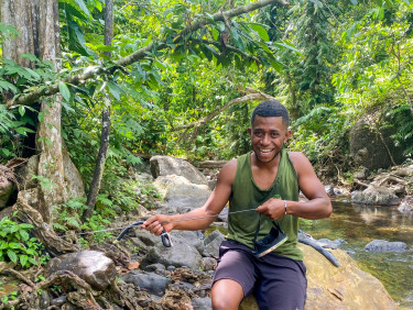 A man in a green vest holds spear-fishing gear by a river in a forest