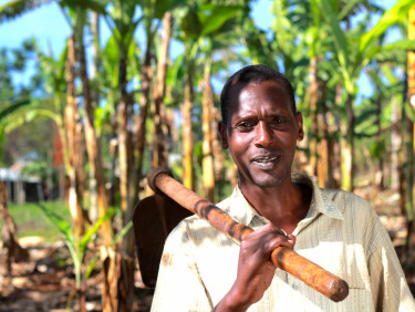 A man in  a white polo shirt with a hoe over his shoulder stands in a lightly forested area