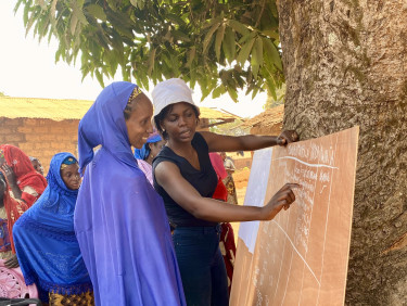 A woman in a hat shows a woman in a hijab a board showing the names and numbers of votes in a committee election