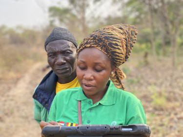 A woman in a headscarf is typing something on an electronic tablet while a man looks over her shoulder