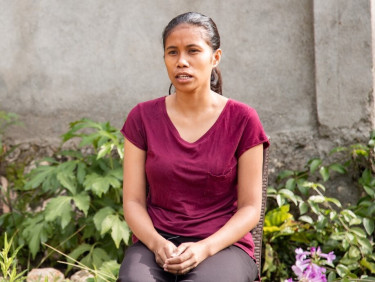 A woman in a fuchsia t-shirt sits in front of a wall and some plants