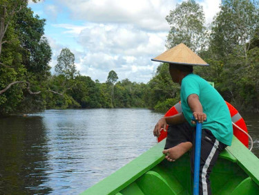A man in a traditional straw hat sits facing away from the camera on the edge of a boat navigating a river with forested banks