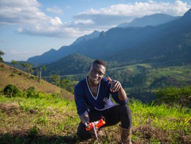 Damian, a forest ambassador, kneeling to measure a seedling with mountains in the background.