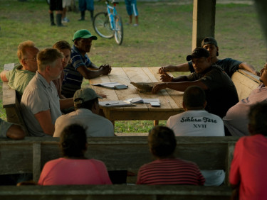 A group of women and men sit around an outdoor table with notebooks and pens on the table
