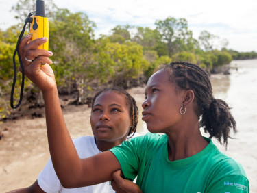Two young women hold up a device on a beach
