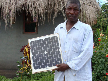 A man in a white shirt holds a solar panel with a hut behind him