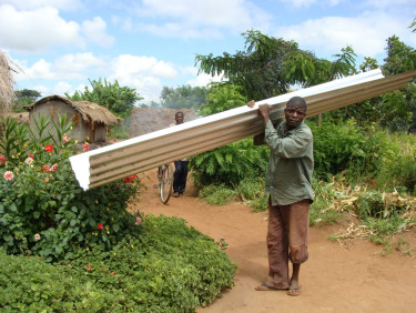 A man in a green shirt holds building materials over his shoulder on a path between plants