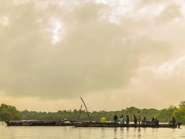 The sea in the foreground with fishing boats with a group of people standing in them in the middle ground and trees and a cloudy sky in the background