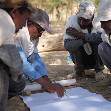 A group of four people crouches over a sheet of paper on the ground and one of them writes on the paper