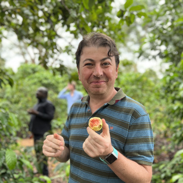 A man in a blue striped polo shirt is smiling and holding a fruit in a forested area