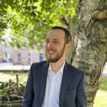 A man in a white shirt and grey jacket stands smiling in front of a tree
