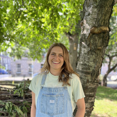 A woman in light denim dungarees stands smiling in front of a tree