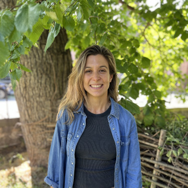 A woman in a blue denim shirt stands smiling in front of a tree