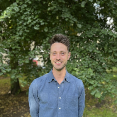 A man in a blue shirt stands smiling in front of a tree