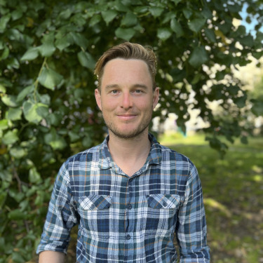 A man in a blue checked shirt stands in front of a tree