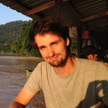 A man in a grey t-shirt smiles outside a wooden building