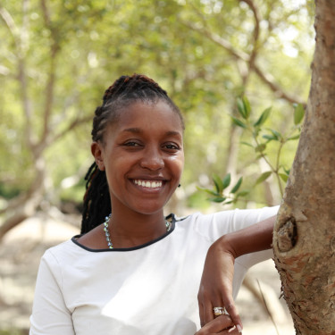 A woman in a white top stands smiling in a forest