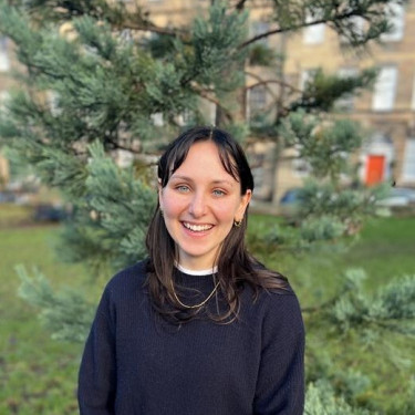 A woman in a black jumper stands smiling in front of a tree