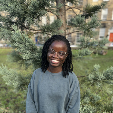 A woman in a grey jumper stands smiling in front of a tree