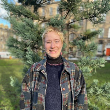 A woman in a patchwork jacket stands smiling in front of a tree