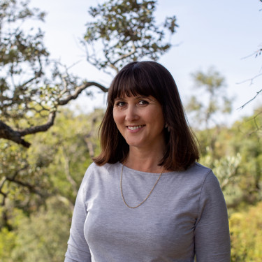 A woman in a white top stands smiling in front of a tree