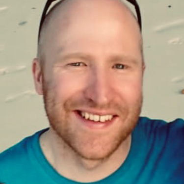 A man in a blue t-shirt smiles at the camera with a pale background