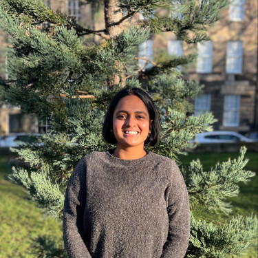A woman in a grey jumper smiles in front of a tree