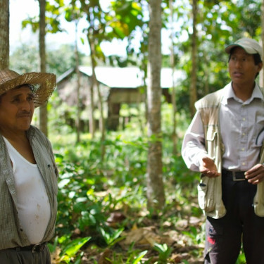 Two men in hats stand talking in a wood with a house in the background