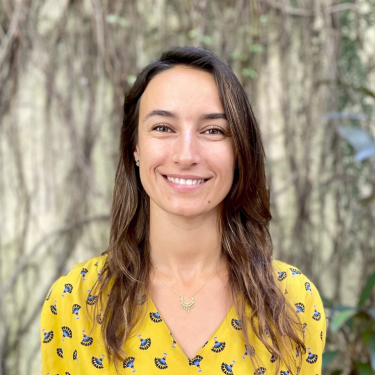 A woman in a yellow top stands smiling in front of a pale background