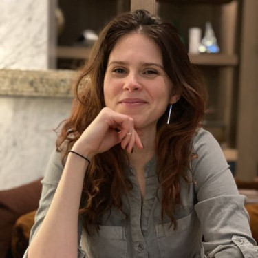 A woman in a khaki shirt sits smiling at a table with a cup of coffee in front of her