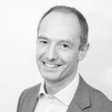 A man in a white shirt and grey jacket smiles at the camera in a greyscale photo