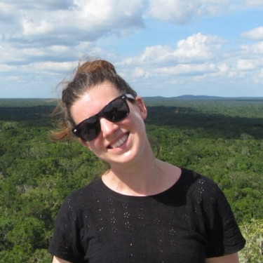 A woman in a black t-shirt stands smiling in front of a rural landscape