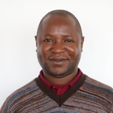 A man in a stripy jumper stands smiling in front of a white wall