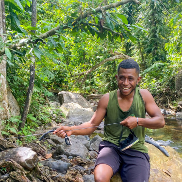 A man in a green vest holds spear-fishing gear by a river in a forest