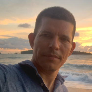 A man in a grey shirt stands on the beach with the sea and a sunset behind him