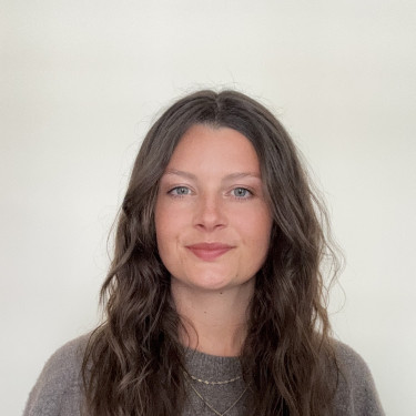 A woman in a grey jumper stands in front of an off-white wall