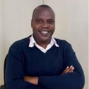 A man in a white shirt and dark blue jumper sits smiling in front of an off-white wall