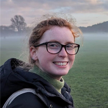 A woman in a black coat stands smiling in front of a misty park