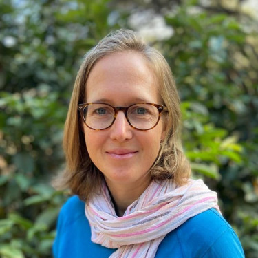 A woman in a blue top and pink scarf stands in front of a tree