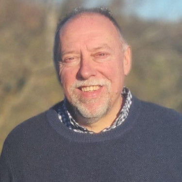 A man in a grey jumper stands smiling in front of a blurred wooded landscape