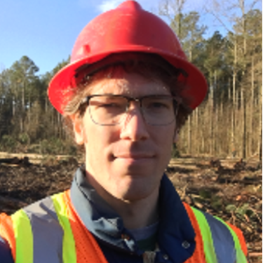 A man in a high-vis vest and an orange hard hat stands in front of a cleared area in a forest