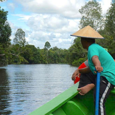A man in a traditional straw hat sits facing away from the camera on the edge of a boat navigating a river with forested banks